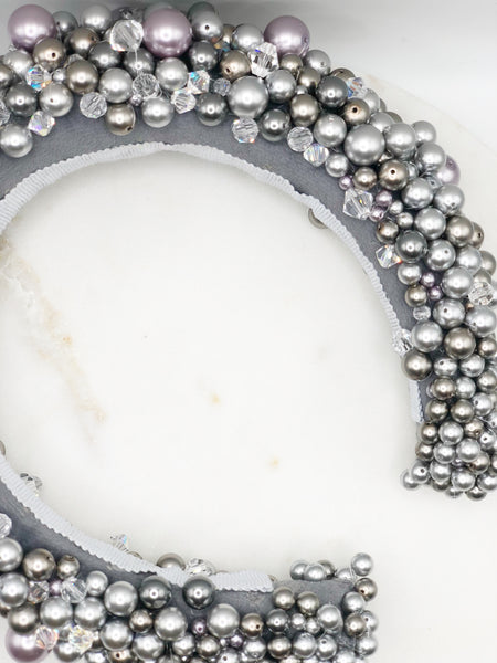 A luxurious grey silk velvet headband heavily adorned with high quality light and dark grey glass pearls and clear crystals of varying sizes displayed on a marble board.