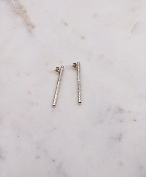 A pair of silver earrings each measuring around 4cm long and lightly textured to resemble that of a twig displayed on a marble board.