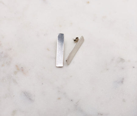 A pair of silver rectangular earrings, one lying on its side, displayed on a marble board.