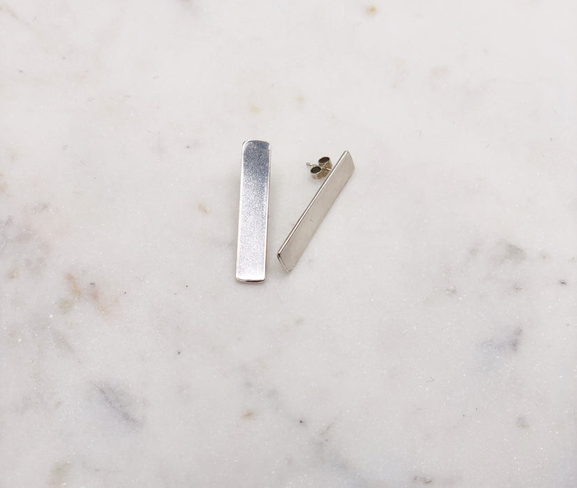 A pair of silver rectangular earrings, one lying on its side, displayed on a marble board.