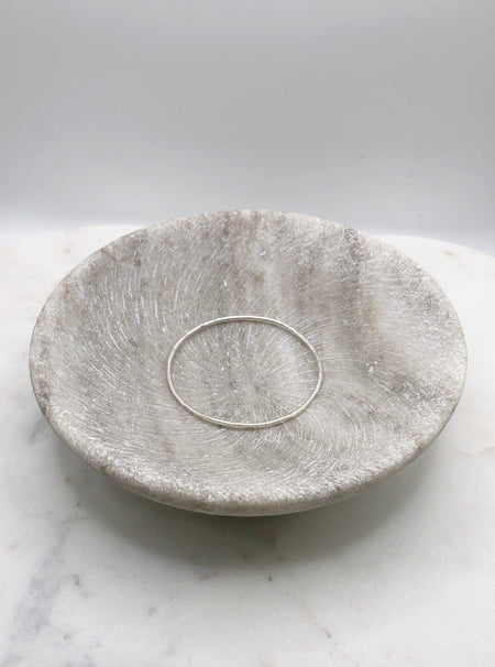 A slim lightly hammered organically shaped circular silver bangle displayed in a marble bowl.