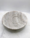 A slim organically shaped circular silver bangle smooth in texture and displayed in a marble bowl.