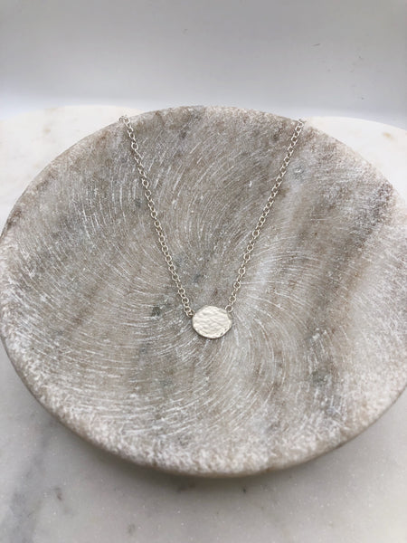 A lightly textured circular silver pendant attached to a medium weight chain displayed in a marble bowl.