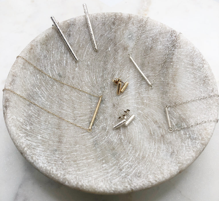 A marble bowl displaying the Twiglet collection. In the centre sits a pair of gold and a pair of silver small twig-like studs each measuring around 1cm in length. Around the edge of the bowl sits a fine gold necklace with a twig-like pendant suspended in the middle of the chain, textured to resemble that of a twig. To the right are the Twiglet earrings in silver measuring around 4cm. Next to these sits the silver version of the Twiglet necklace and finally there is the Twiglet bracelet in silver.