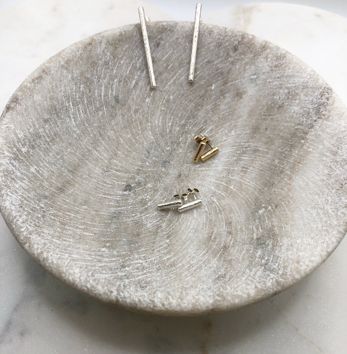 A marble bowl displaying three pairs of earrings. In the centre of the bowl sits a pair of gold and a pair of silver small twig-like studs. These measure around 1cm long and are lightly textured to resemble that of a twig. Draped on the edge of the bowl above theses studs sits a pair of silver earrings. These measure around 4cm long and are lightly textured to resemble that of a twig.