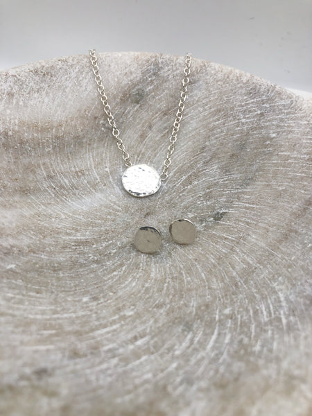 A lightly textured circular silver pendant attached to a medium weight chain and a pair of small silver studs in the same circular and textured design. Both displayed in a marble bowl.