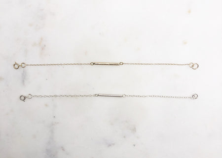 Two bracelets, one gold and one silver displayed on a marble board. Both bracelets feature a textured “twig” bar suspended in the middle of a fine chain