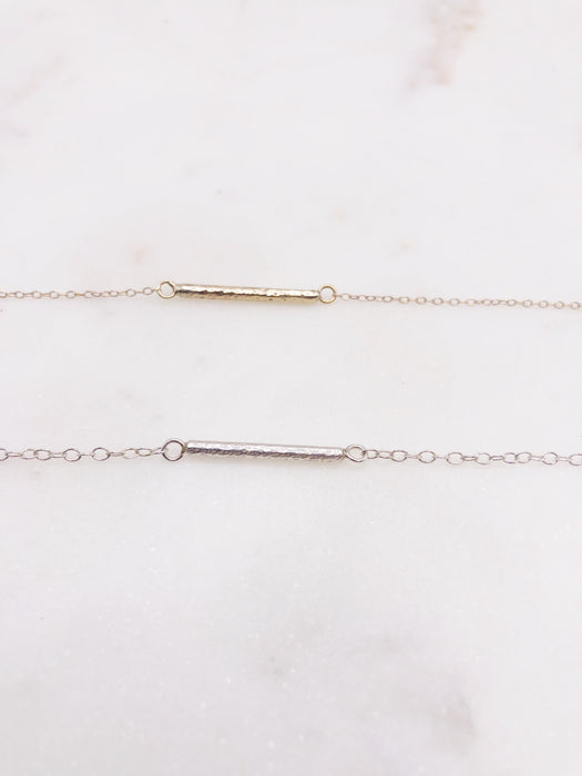 A close up photograph of two bracelets, one gold and one silver displayed on a marble board. Both bracelets feature a textured “twig” bar suspended in the middle of a fine chain. 