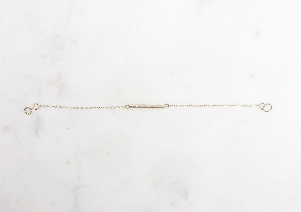 A delicate gold chain bracelet with a decorative solid gold “twig” bar suspended in the middle of the chain and displayed on a marble board.