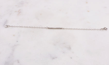 A delicate silver chain bracelet with a decorative solid silver “twig” bar suspended in the middle of the chain and displayed on a marble board.