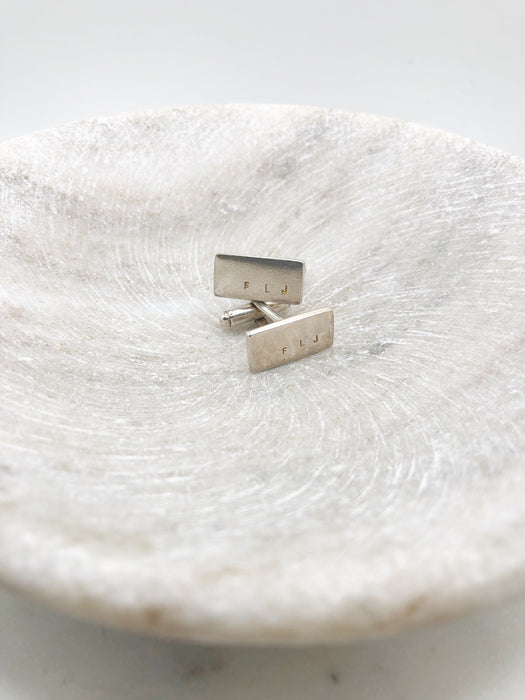 A pair of silver rectangle cufflinks with the letters F L J engraved on both cufflinks displayed in a shallow marble bowl.