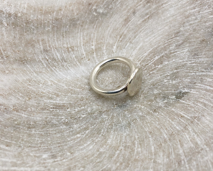 A chunky silver ring consisting of a 3mm silver wire band with a lightly textured circular silver nugget/pebble attached. Sat in a marble bowl.