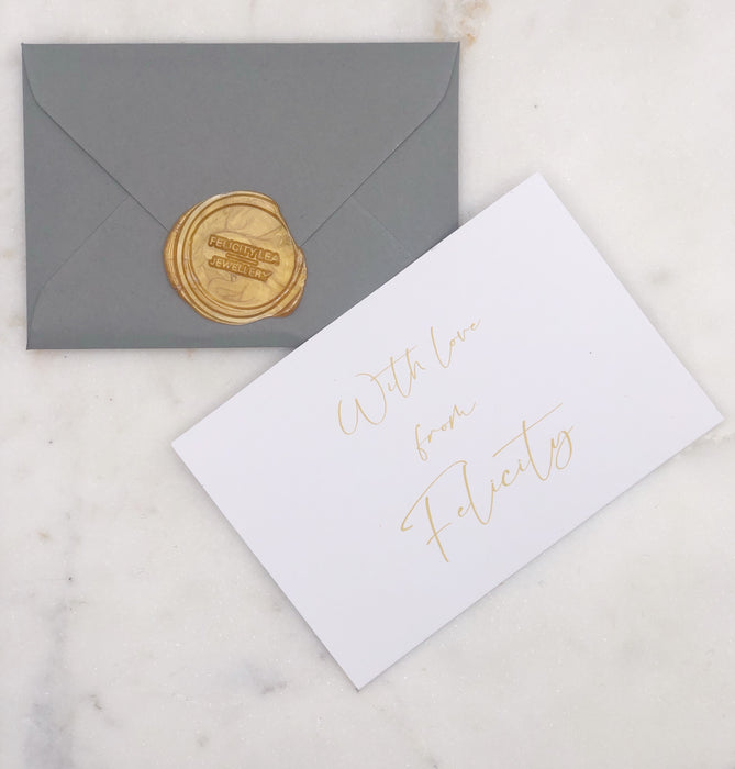 A small grey envelope with a gold Felicity Lea Jewellery logo wax seal and a white card with the words “With love from Felicity” written in gold calligraphy. Both items displayed on a marble board.