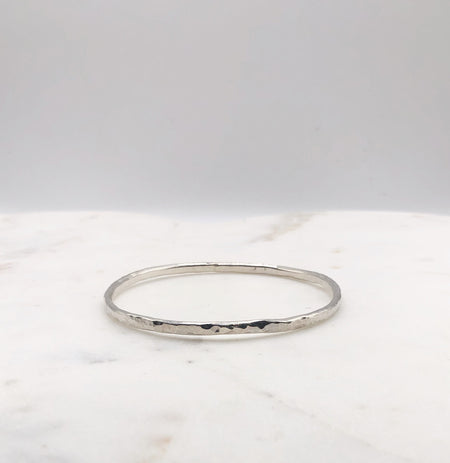 A lightly hammered organically shaped circular silver bangle displayed on a marble board.