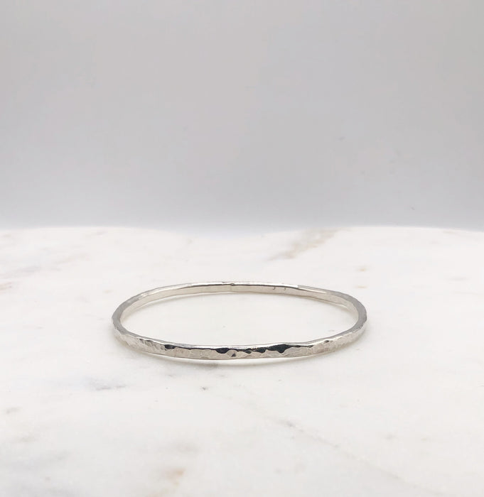 A lightly hammered organically shaped circular silver bangle displayed on a marble board.