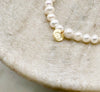 A close up of a white pearl bracelet made from Fresh Water Pearls with a circular gold tag engraved with the letters FLJ displayed on a marble bowl