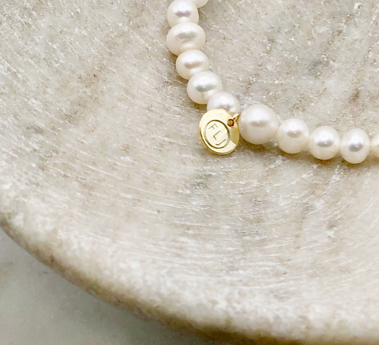 A close up of a white pearl bracelet made from Fresh Water Pearls with a circular gold tag engraved with the letters FLJ displayed on a marble bowl