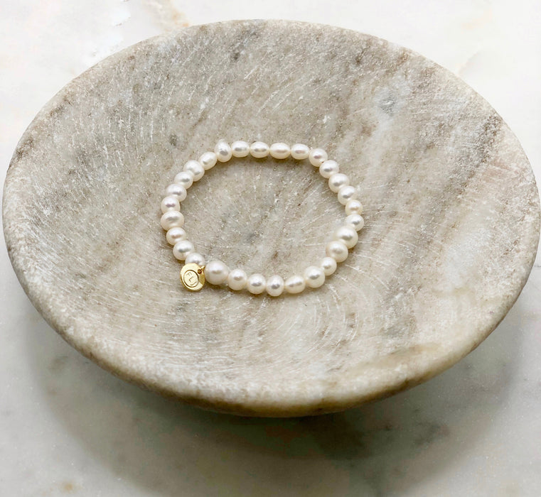 A white pearl bracelet made from Fresh Water Pearls with a circular gold tag engraved with the letters FLJ displayed on a marble bowl.