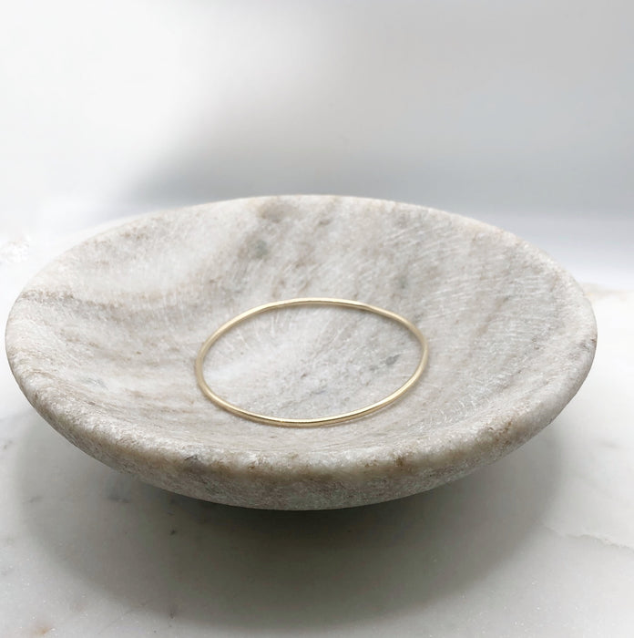 A slim organically shaped circular gold bangle smooth in texture and displayed in a marble bowl.